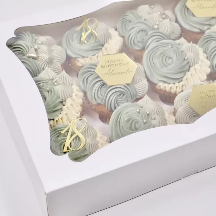 Zwölf Cupcakes mit hellblauer Creme und goldenen "Happy Birthday" Auflegern in einer weißen Schachtel