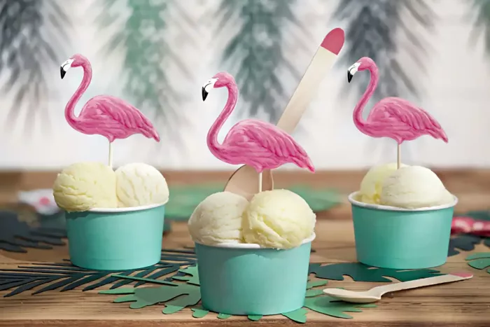 Drei Eisbecher mit Eis dekoriert mit drei Flamingo Cake Toppern vor weißem Hintergrund mit Pflanzen