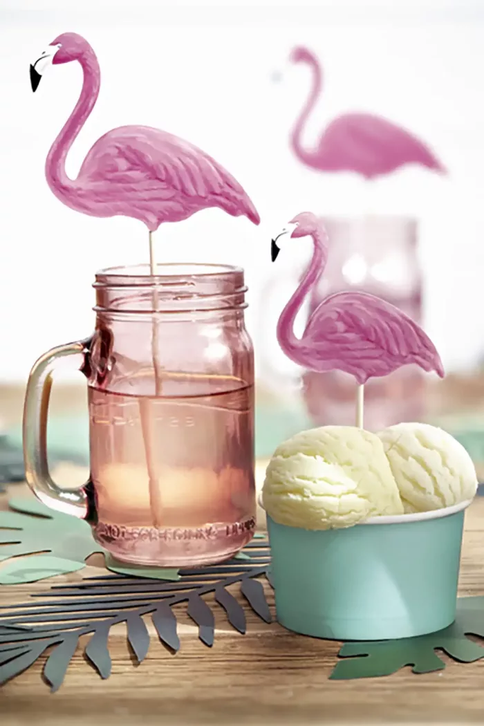 Ein Eisbecher und ein gefülltes Glas dekoriert mit zwei Flamingo Cake Toppern auf einem Holztisch