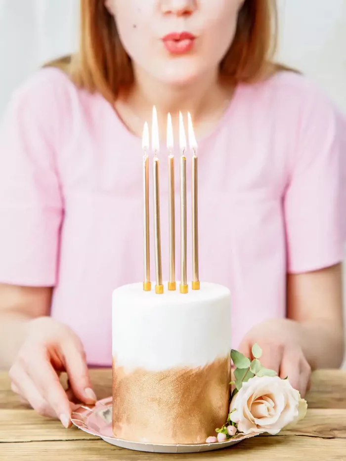 Eine weiß-goldene Torte mit brennenden, goldenen Kerzen mit einer Frau, die sie ausblasen möchte, im Hintergrund