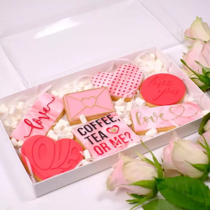 Eine weiße Sweet Box mit transparentem Deckel, Keksen mit verschiedenen Prägungen in Rosa und Rot zum Valentinstag, sowie Mini Marshmallows