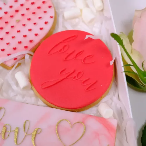 Ein runder Keks mit rotem Zuckerguss und gestempelter Prägung "love you", liegend in einer Box mit anderen Keksen zum Valentinstag und Mini Marshmallows