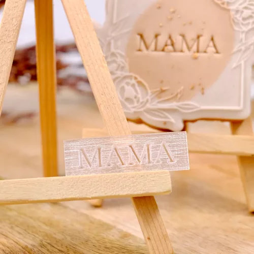 Ein rechteckiger Keksstempel aus transparentem Acryl mit dem Schriftzug "Mama" auf einer kleinen Staffelei