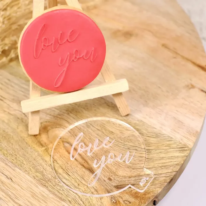 Ein runder Keks mit rotem Zuckerguss und gestempelter Prägung "love you" auf einer kleinen Staffelei, davor liegend ein Keksstempel aus transparentem Acrylglas