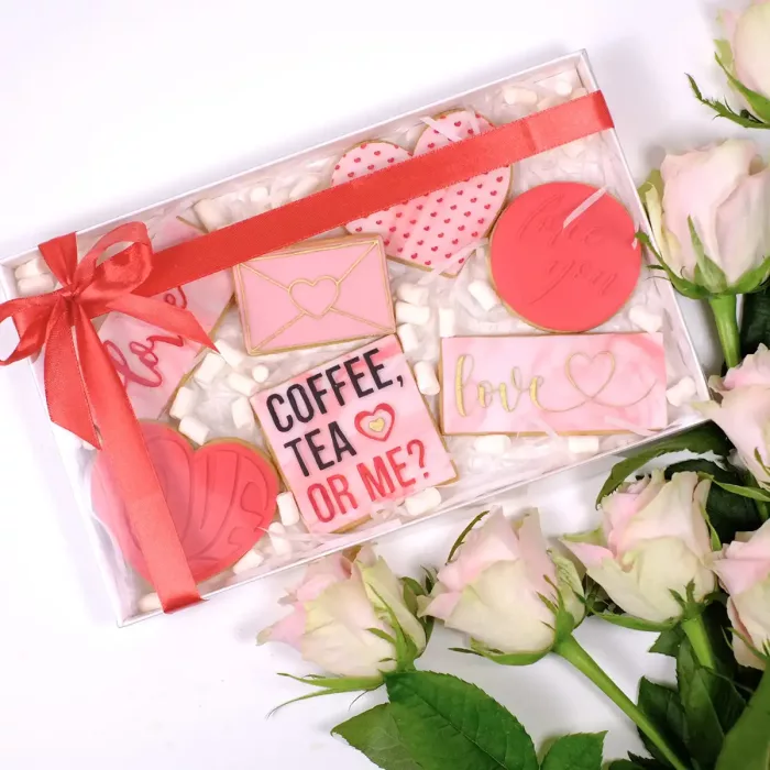 Eine weiße Sweet Box mit transparentem Deckel, rotem Band mit Schleife, Keksen mit verschiedenen Prägungen in Rosa und Rot zum Valentinstag, sowie Mini Marshmallows