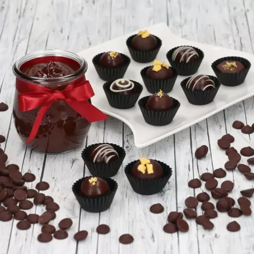 Ein weißer Teller mit mehreren dunkle Zartbitter Pralinen in schwarzen Förmchen, davor weiße Schokoladendrops und daneben ein Glas mit dunlem Schokopudding