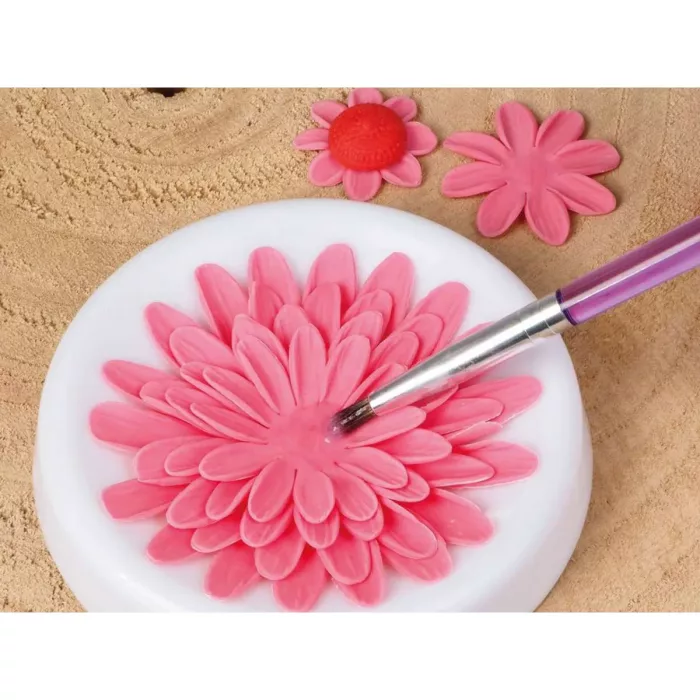 Eine pinke Blume aus Fondant auf einem weißen Podest aus Fondant, auf das mit einem Pinsel transparenter, essbarer Kleber aufgetragen wird