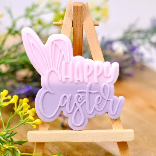 Ein pastellfarbener Keks in Rosa und Lila mit dem gestempelten Schriftzug "Happy Easter" und Hasenohren auf einer kleinen Staffelei