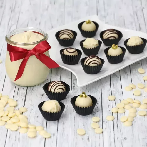 Ein weißer Teller mit mehreren weißen Pralinen in schwarzen Förmchen, davor weiße Schokoladendrops und daneben ein Glas mit weißem Pudding