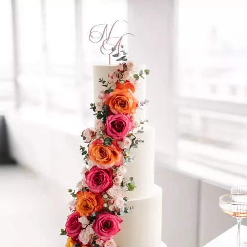 Eine dreistöckige weiße Torte mit Rosen und einem Cake Topper aus Acryl in Rosegold mit den Initialen "N" und "A" in geschwungener Schrift