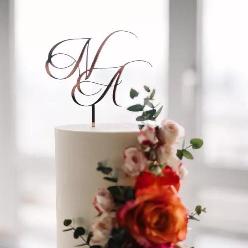 Nahaufnahme eines Cake Toppers aus Acryl in Rosegold mit den Initialen "N" und "A" in geschwungener Schrift auf weißer Torte mit orangenen Blumen