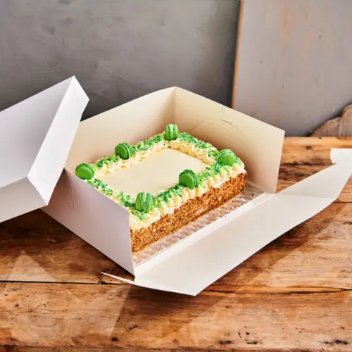 Aufgeklappte weiße Tortenbox mit beige-grüner Torte darin, auf einem Holztisch stehend