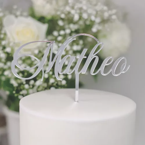 Nahaufnahme eines Cake Toppers aus Silber Acryl mit dem Schriftzug "Matheo" auf einer weißen, dekorierten Torte