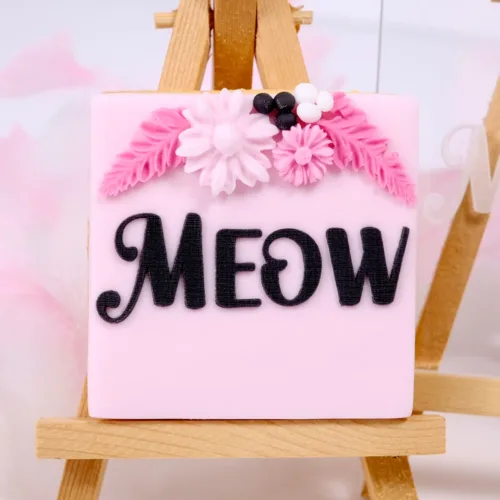 Schwarz-rosa Keks mit pinken Details mit "Meow" Schriftzug auf einer Staffelei.