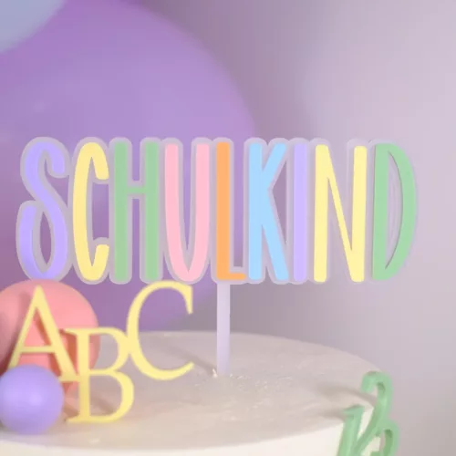 Weiße Torte mit bunten Dekoelementen und einem Cake Topper in Pastellfarben mit der Aufschrift ‚Schulkind‘, verziert mit ABC- und Zahlenmotiven 123. Einschulung