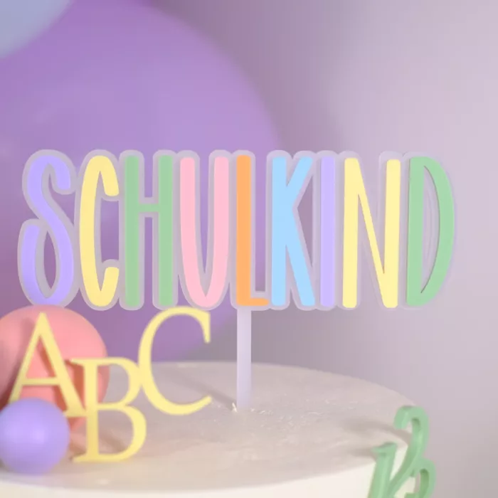 Weiße Torte mit bunten Dekoelementen und einem Cake Topper in Pastellfarben mit der Aufschrift ‚Schulkind‘, verziert mit ABC- und Zahlenmotiven 123. Einschulung