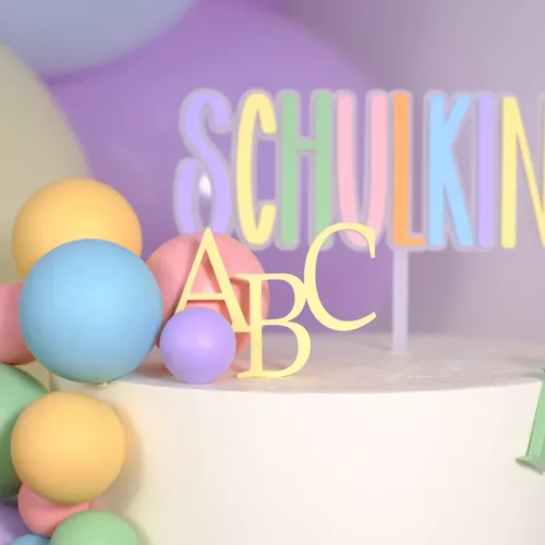 Weiße Torte mit bunten Dekoelementen und einem Cake Topper in Pastellfarben mit der Aufschrift ‚Schulkind‘, verziert mit ABC- und Zahlenmotiven 123. Einschulung