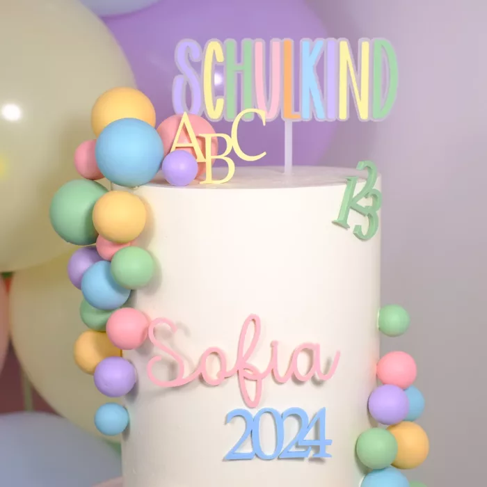 Weiße Torte mit bunten Dekoelementen und einem Cake Topper in Pastellfarben mit der Aufschrift ‚Schulkind‘, verziert mit ABC- und Zahlenmotiven 123. Einschulung