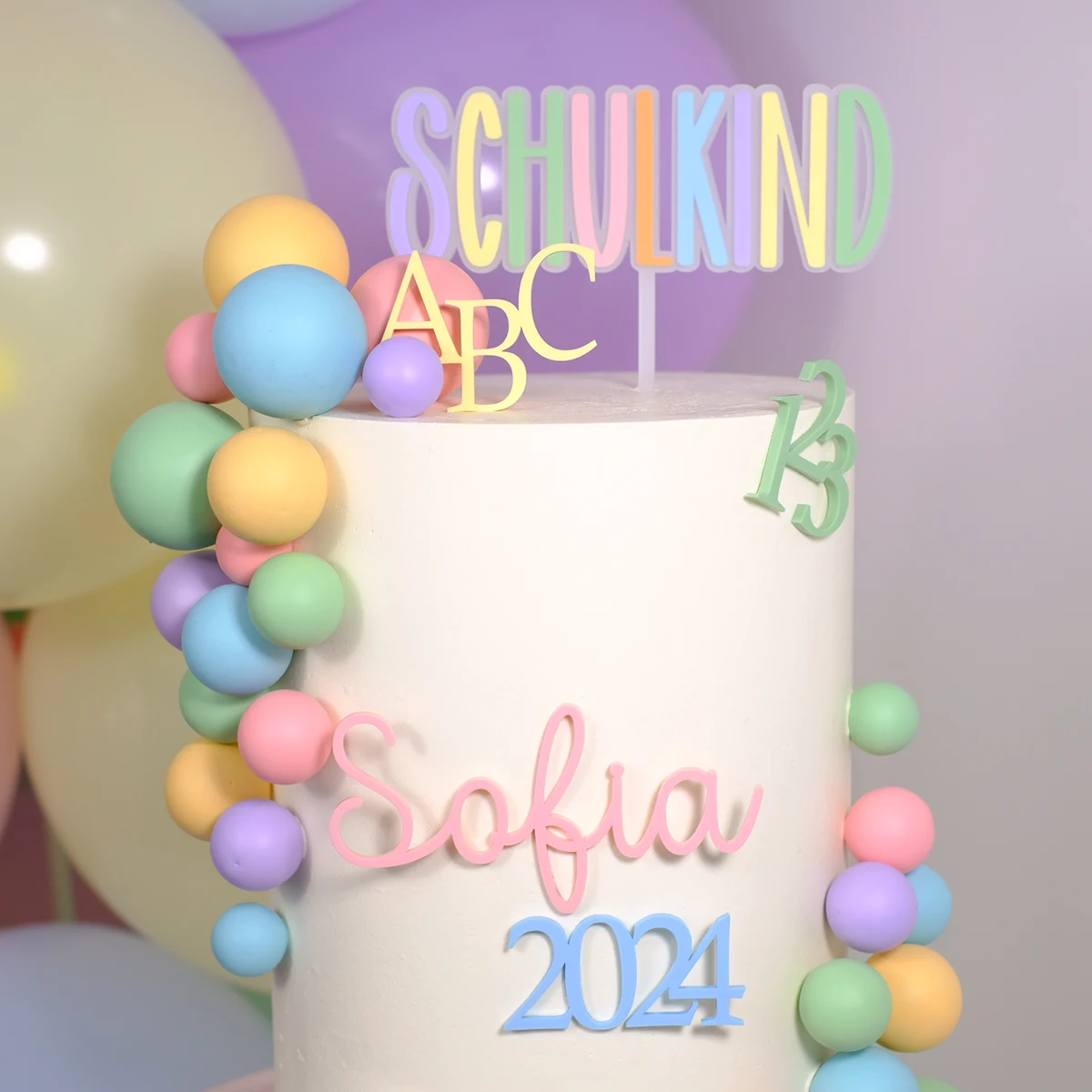 Weiße Torte mit bunten Dekoelementen und einem Cake Topper in Pastellfarben mit der Aufschrift ‚Schulkind‘, verziert mit ABC- und Zahlenmotiven 123. Einschulung
