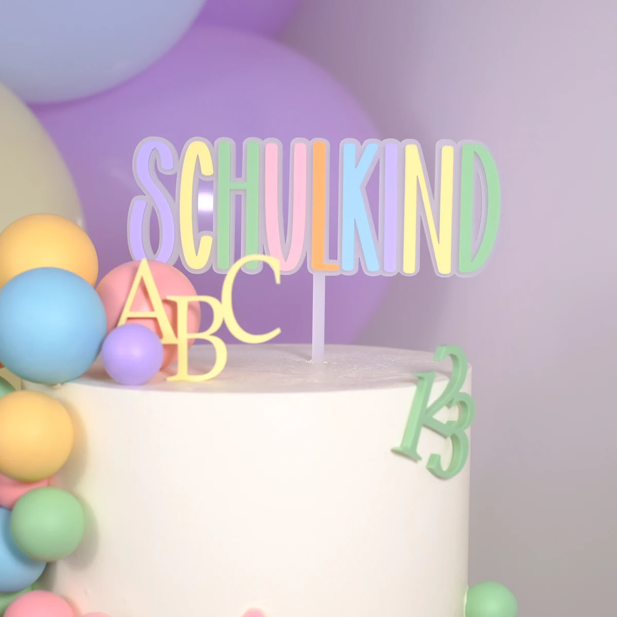 Weiße Torte mit bunten Dekoelementen und einem Cake Topper in Pastellfarben mit der Aufschrift ‚Schulkind‘, verziert mit ABC- und Zahlenmotiven 123. Einschulung