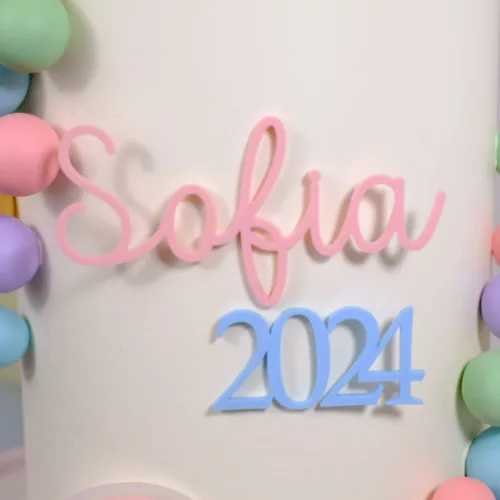 Ein bunter Cake Topper ohne Stiel mit personalisierten Namen in zartem Rosa und der Jahreszahl 2024 in Hellblau schmückt eine weiße Torte. Umrahmt wird die Deko von pastellfarbenen Kugeln in Gelb, Grün, Rosa, Lila und Blau. Am unteren Rand der Torte befindet sich ein kleiner Regenbogen in sanften Farben mit einem grünen Herz in der Mitte.