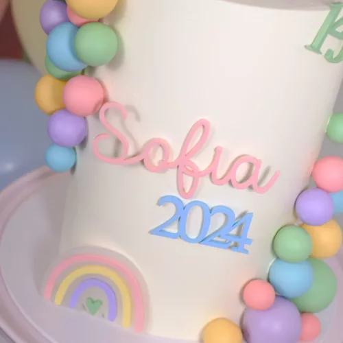 Ein bunter Cake Topper ohne Stiel mit personalisierten Namen in zartem Rosa und der Jahreszahl 2024 in Hellblau schmückt eine weiße Torte. Umrahmt wird die Deko von pastellfarbenen Kugeln in Gelb, Grün, Rosa, Lila und Blau. Am unteren Rand der Torte befindet sich ein kleiner Regenbogen in sanften Farben mit einem grünen Herz in der Mitte.