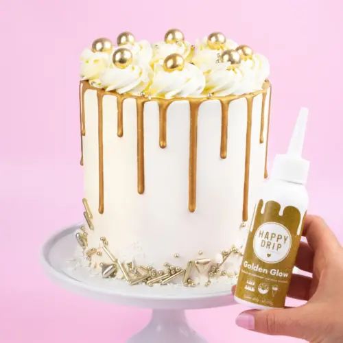 Weiße Spritzflasche mit goldenem Happy Sprinkles Happy Drip auf rosa Hintergrund mit einer Torte