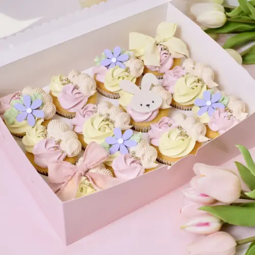 Mehrere Cupcakes mit rosa-gelber Buttercreme, Blumen und Hasen Toppern und Sprinkles in einer rosanen Cupcake Box