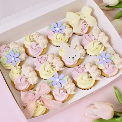 Mehrere Cupcakes mit rosa-gelber Buttercreme, Blumen und Hasen Toppern und Sprinkles in einer rosanen Cupcake Box