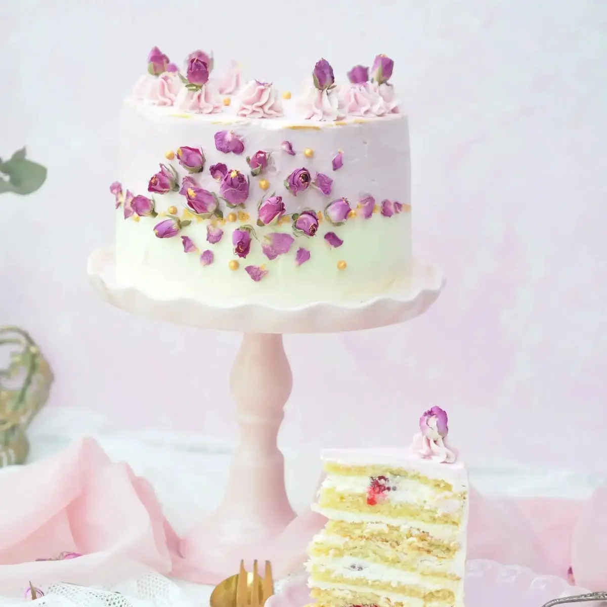 Pastellfarbene Torte mit getrockneten Damaszener-Rosenknospen, rosa Cremetupfen und goldenen Zuckerperlen auf einer Tortenplatte.