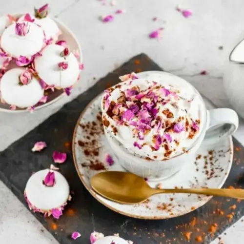 Weiße Kaffeetasse mit Sahnehaube und lila-rosa Rosenblättern, daneben weiß glasiertes Gebäck mit Rosenknospen.